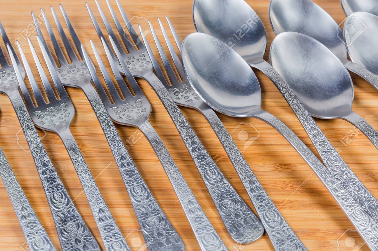 Stainless steel eating utensils consisting of spoons and forks laid out on the wooden surface, fragment close-up in selective focus