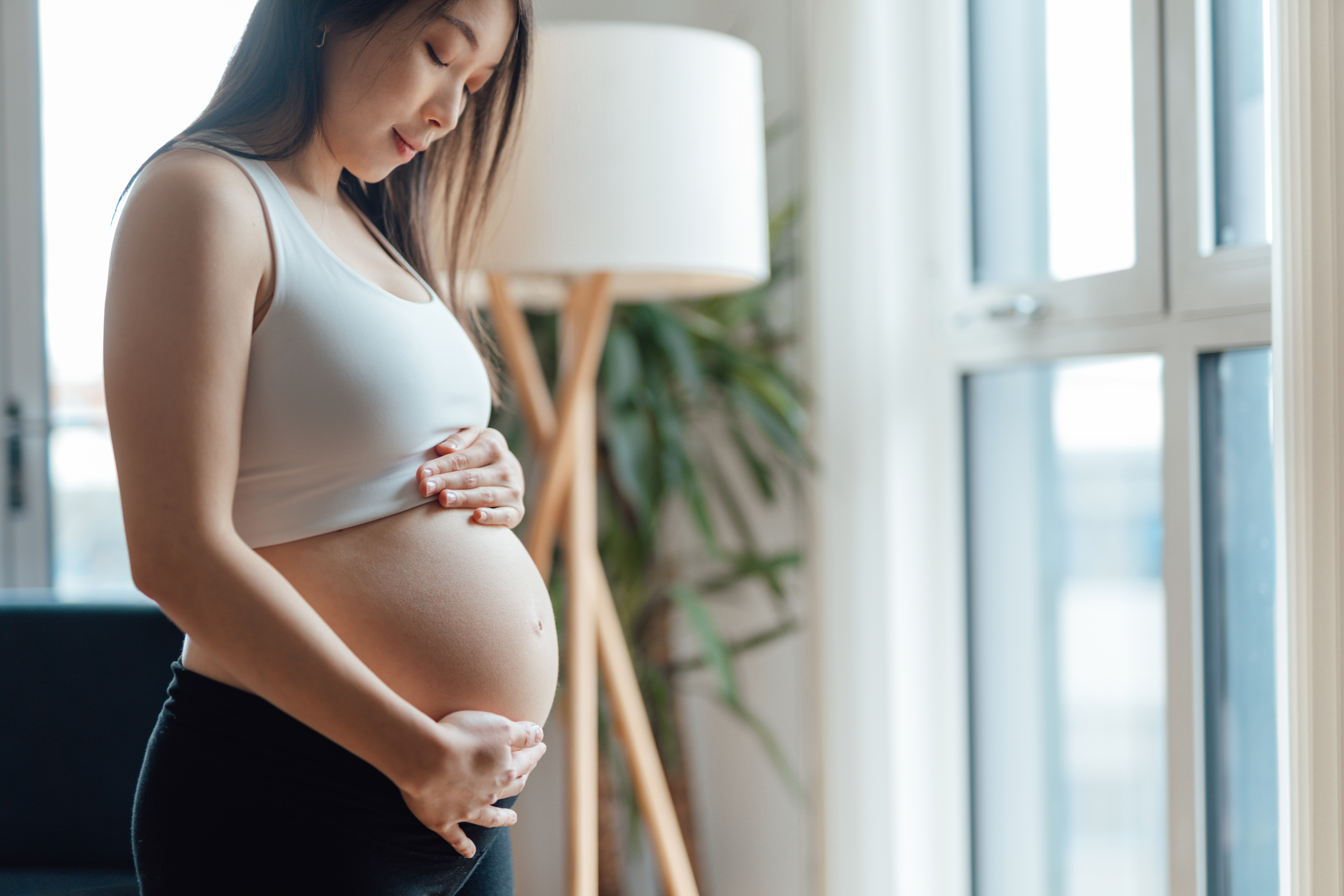 Young Asian pregnant woman looking down and holding her bump thoughtfully by window in modern living room on a sunny day.