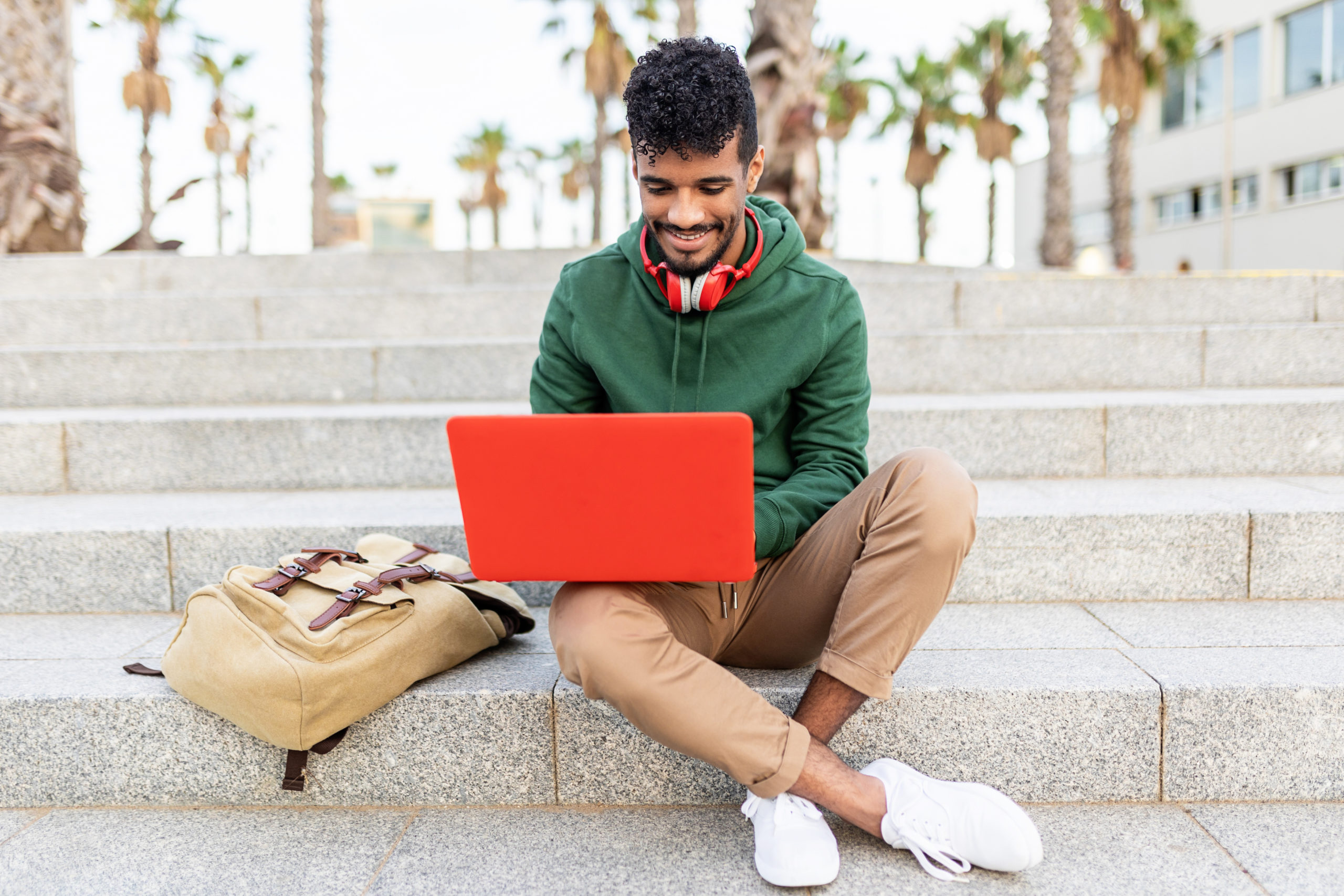 Young hispanic man sitting outdoors and working on laptop