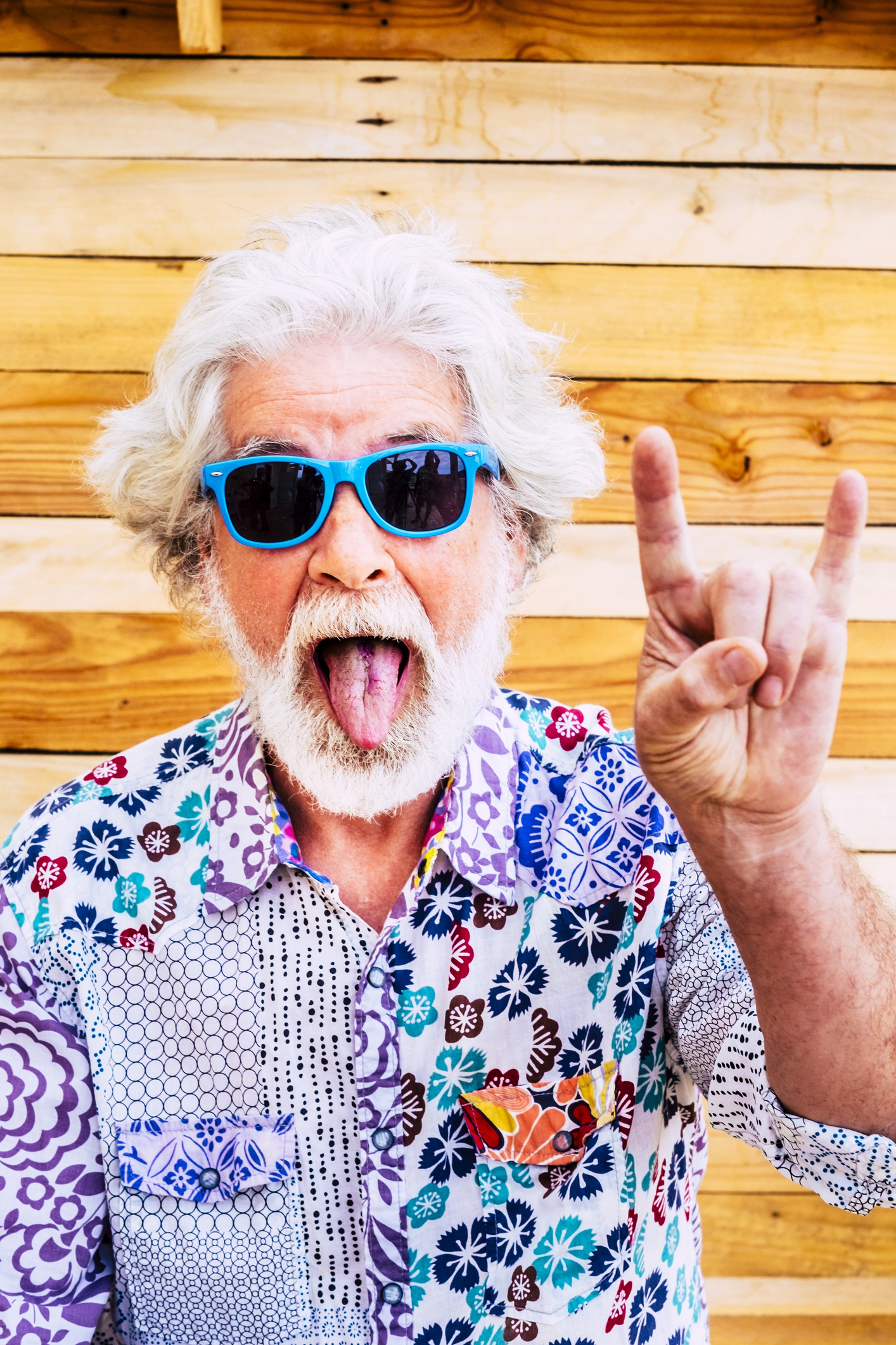 Senior man making funny expression and gesturing against wooden wall. Portrait of elderly man in funky shirt and sunglasses. Old man gesturing with hands and making humorous face