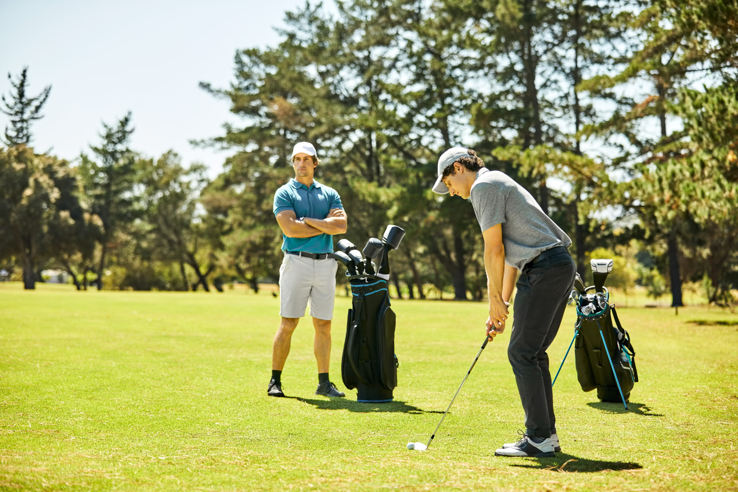 Side view of male golfer playing on field during sunny day. Young man is taking shot with golf club while friend standing in background. They are in sportswear.