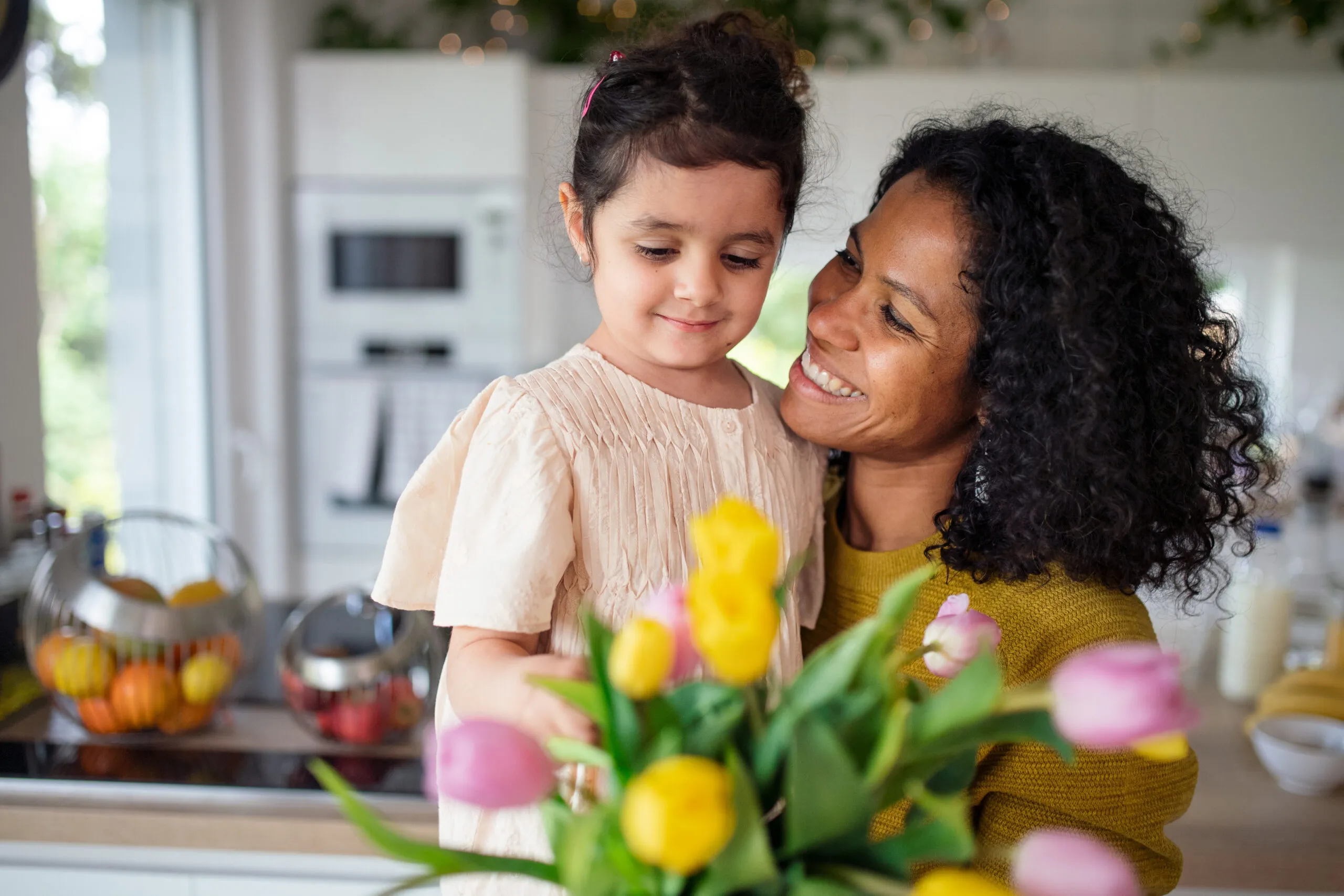Multiracial girl giving flowers her mother in their kitchen, having fun.