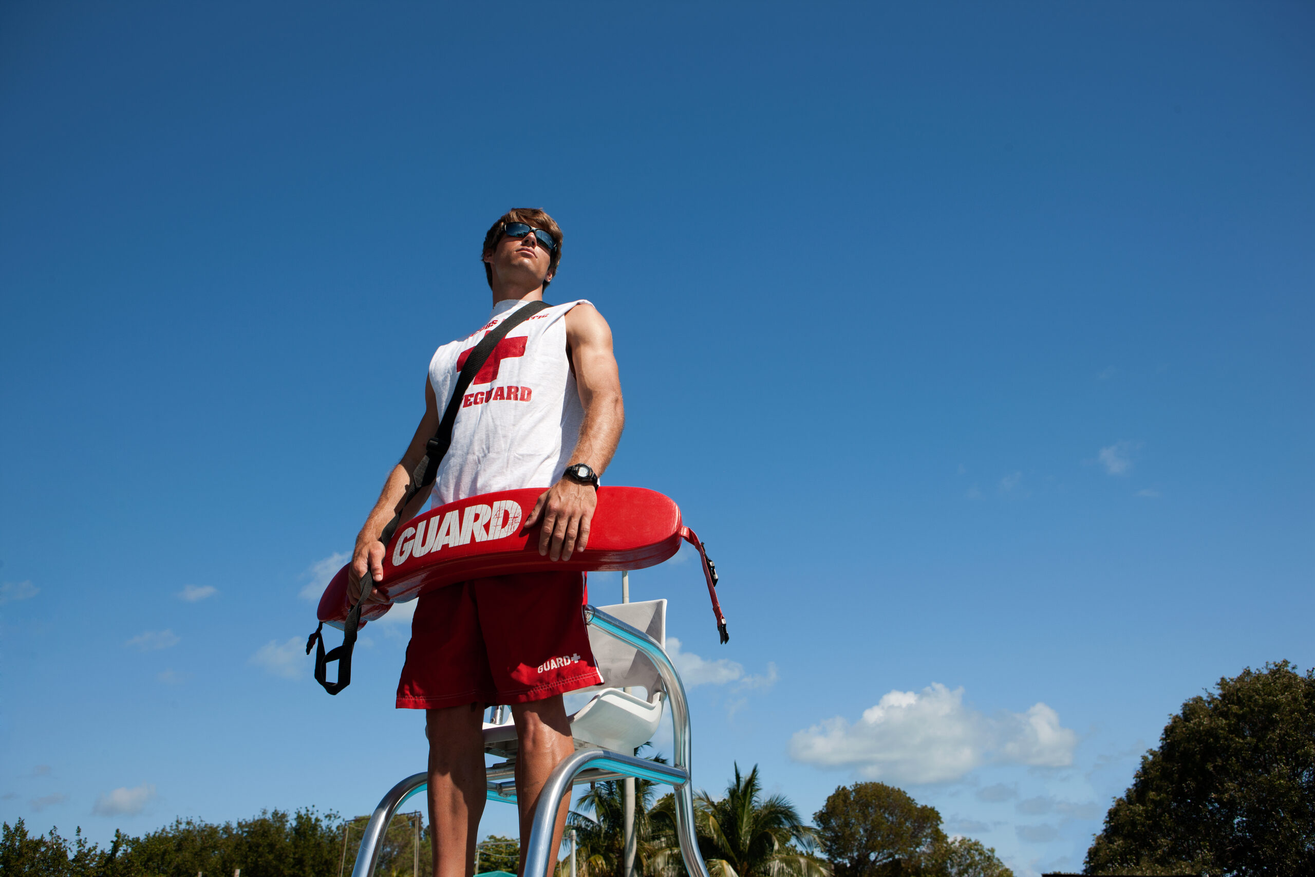 21 year old male, posing as a lifeguard at Jacobs Aquatic Center, wearing lifeguard shirt and  holding lifeguard's rescue buoy.