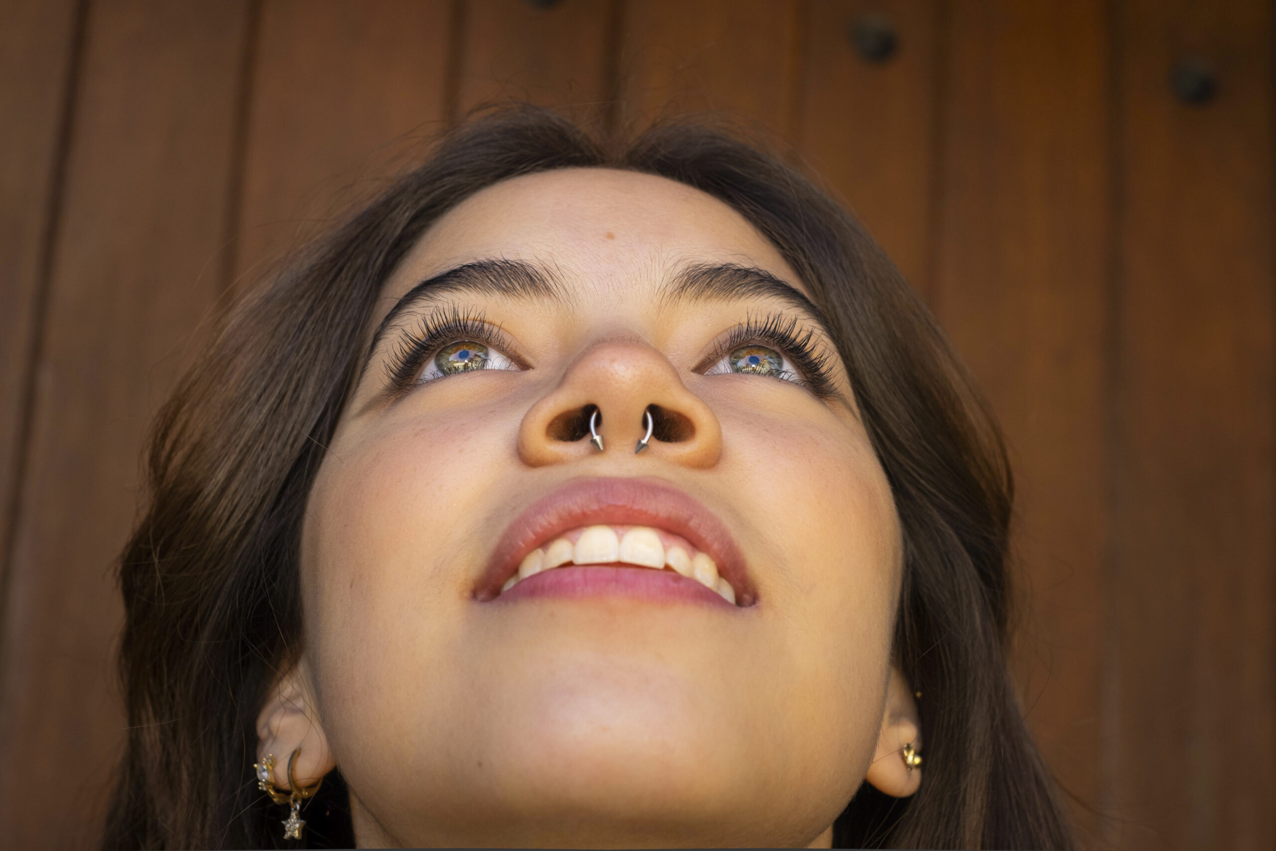 Young Latino woman looking up and showing a nose ring piercing, brown wooden background