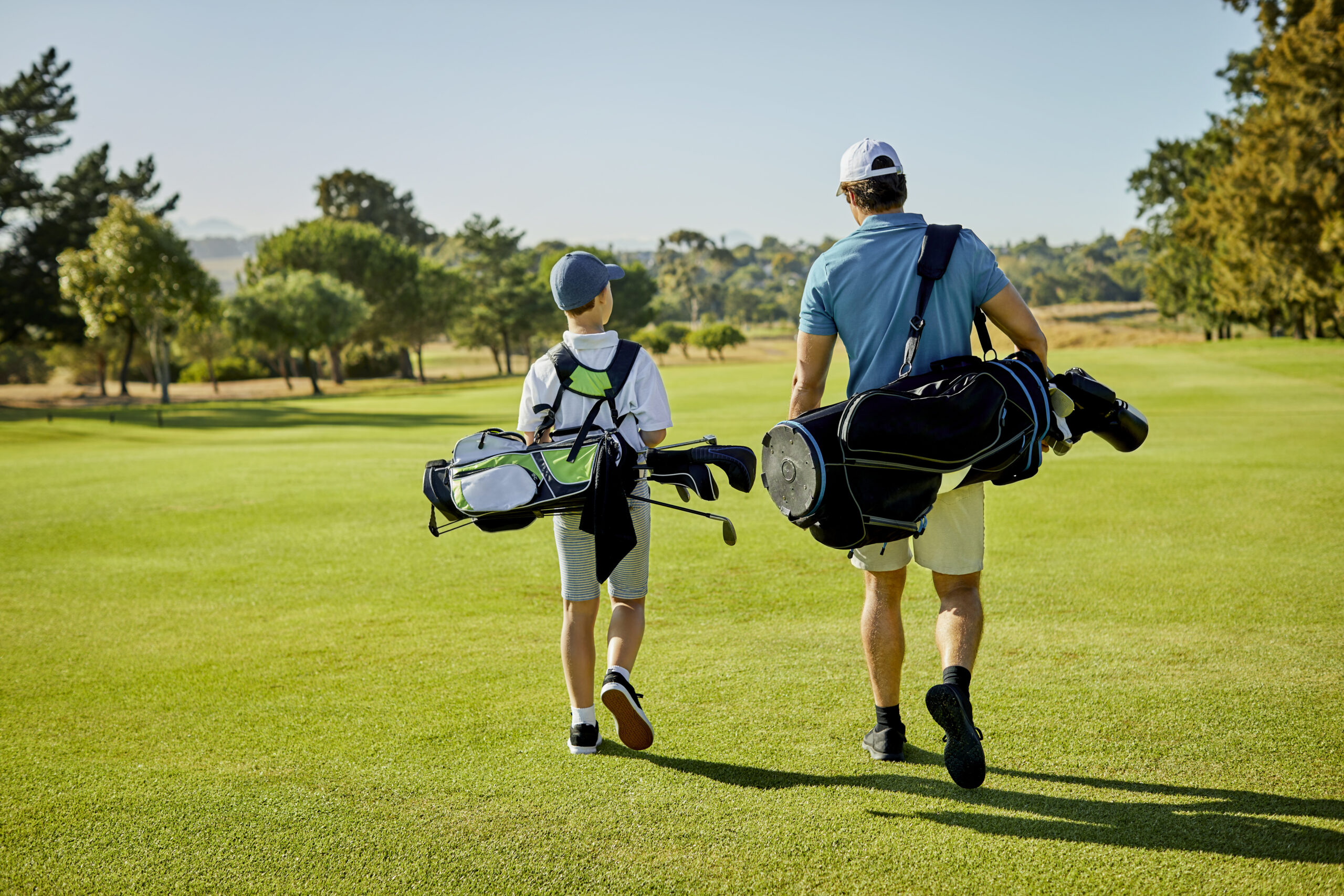 Rear view of man walking with boy on golf course. Father and son are carrying bags at sports field. They are in sportswear.