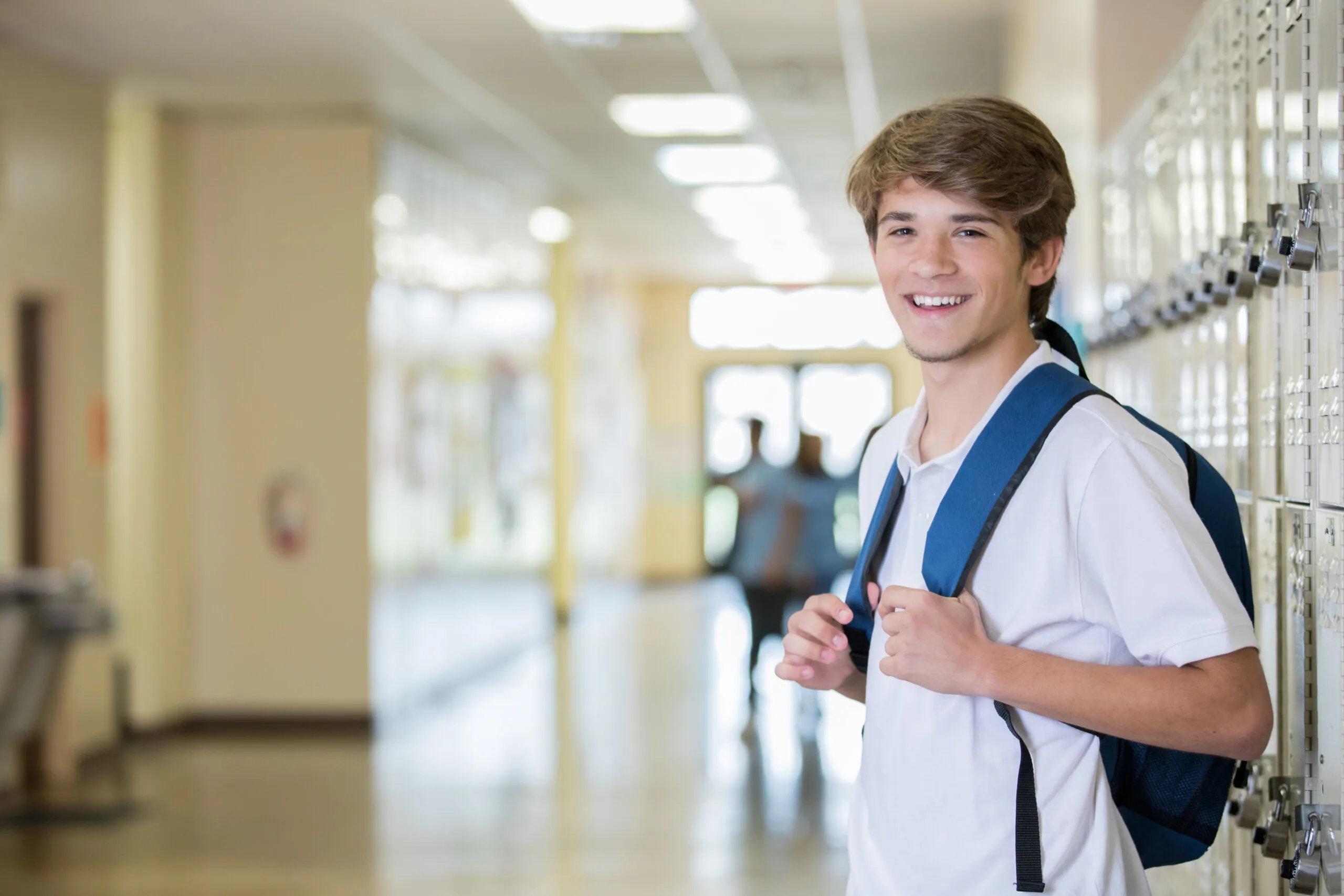 High school senior boy smiles while standing near locker and wearing backpack