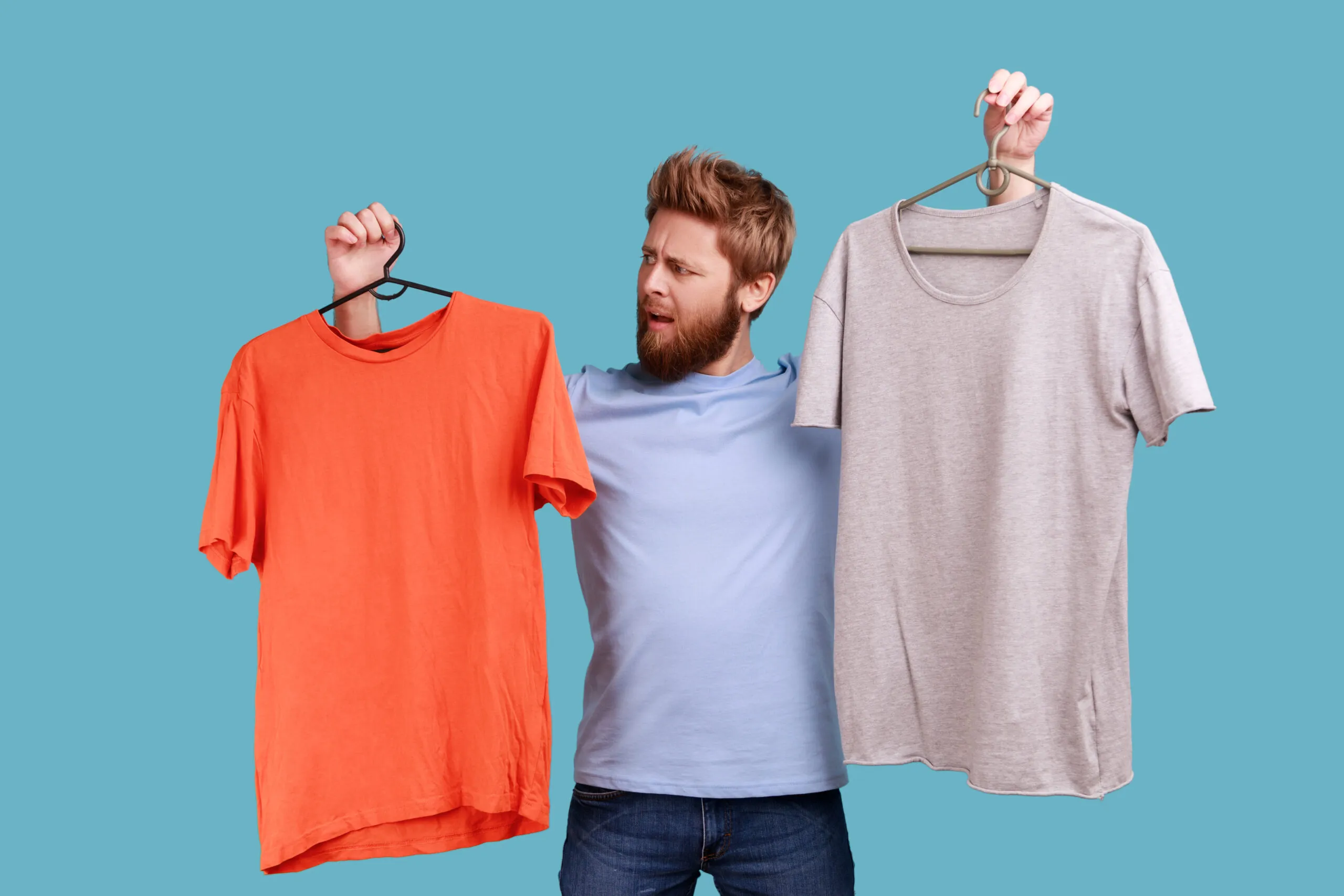 Man frowning face holding two hangers with gray and orange T-shirts, hard to choose the best attire.